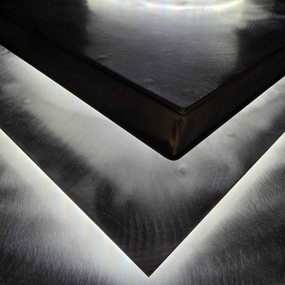 Détail de finition de l’applique Sype 011, surface d’acier brossé et reflets de lumière subtilement diffusés.